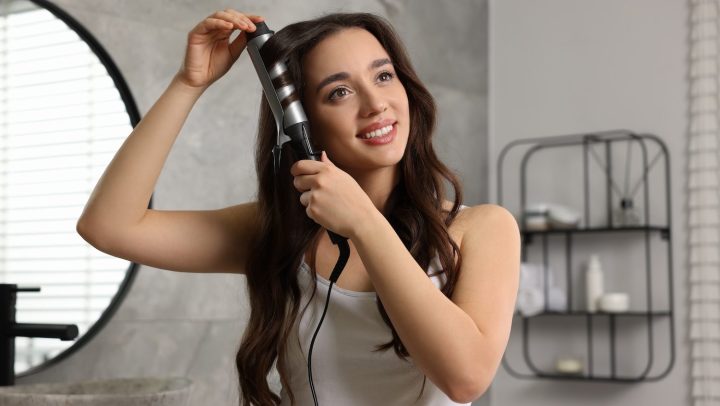 Mujer sonriente se peina frente al espejo usando una tenaza.