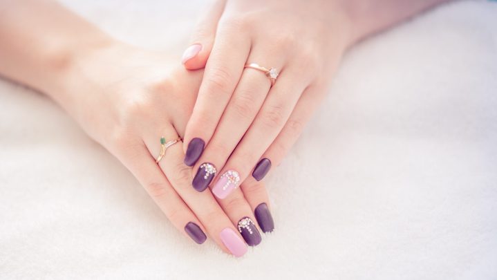 Primer plano de una manicura en color beige y morado con pedrería en algunas uñas.