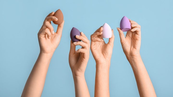 Manos femeninas con diferentes esponjas de maquillaje sobre fondo azul.