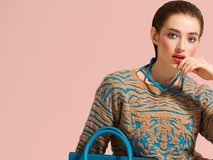 Modelo posa con ropa de diseño y bolso azul sobre fondo de color claro.