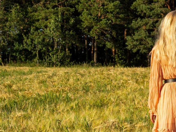 Mujer joven de pie en un campo de trigo con la salida del sol al fondo.