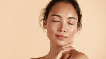 Hermosa modelo asiática tocando piel facial hidratada brillante y fresca sobre fondo beige de cerca.