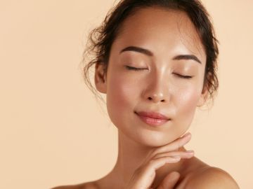 Hermosa modelo asiática tocando piel facial hidratada brillante y fresca sobre fondo beige de cerca.